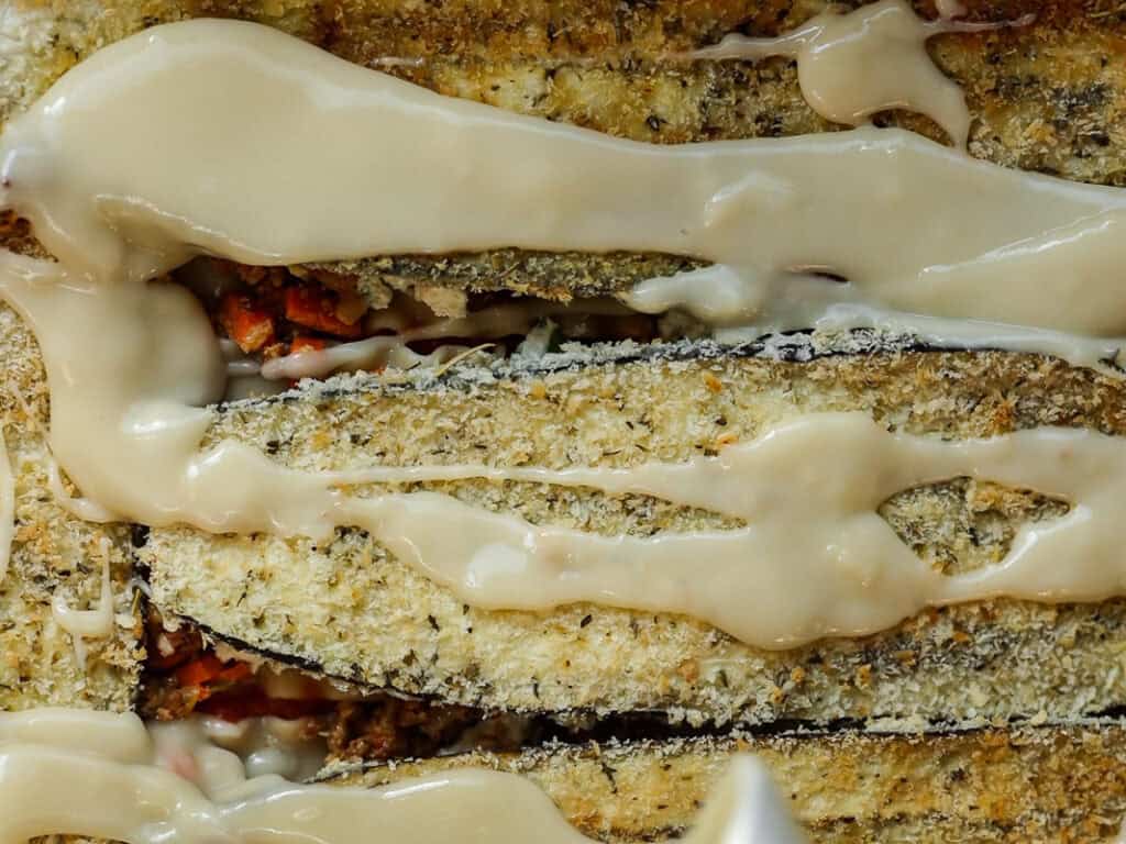 Close-up of layered breaded eggplant slices drizzled with a creamy white sauce, revealing bits of red and green underneath. The texture of the eggplants is crispy, and the sauce adds a glossy sheen.